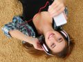 Beautiful blonde smiling woman lying on carpet floor wearing headphones, holding cell phone and listening music portrait. Modern urban life, audio book, radio or education concept